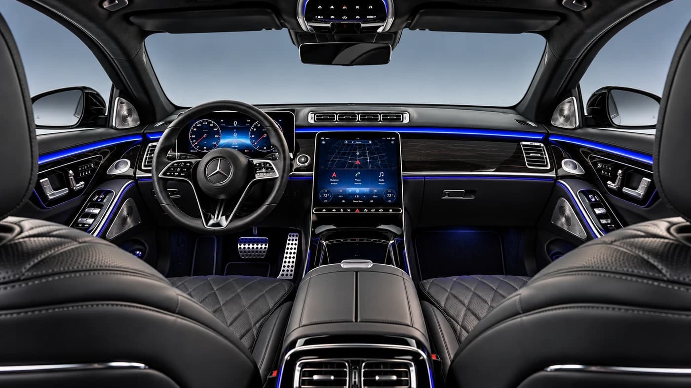 Mercedes-Benz S-Class cabin from the driver's side: black leather seats, blue lighting, a center screen and wood trim.