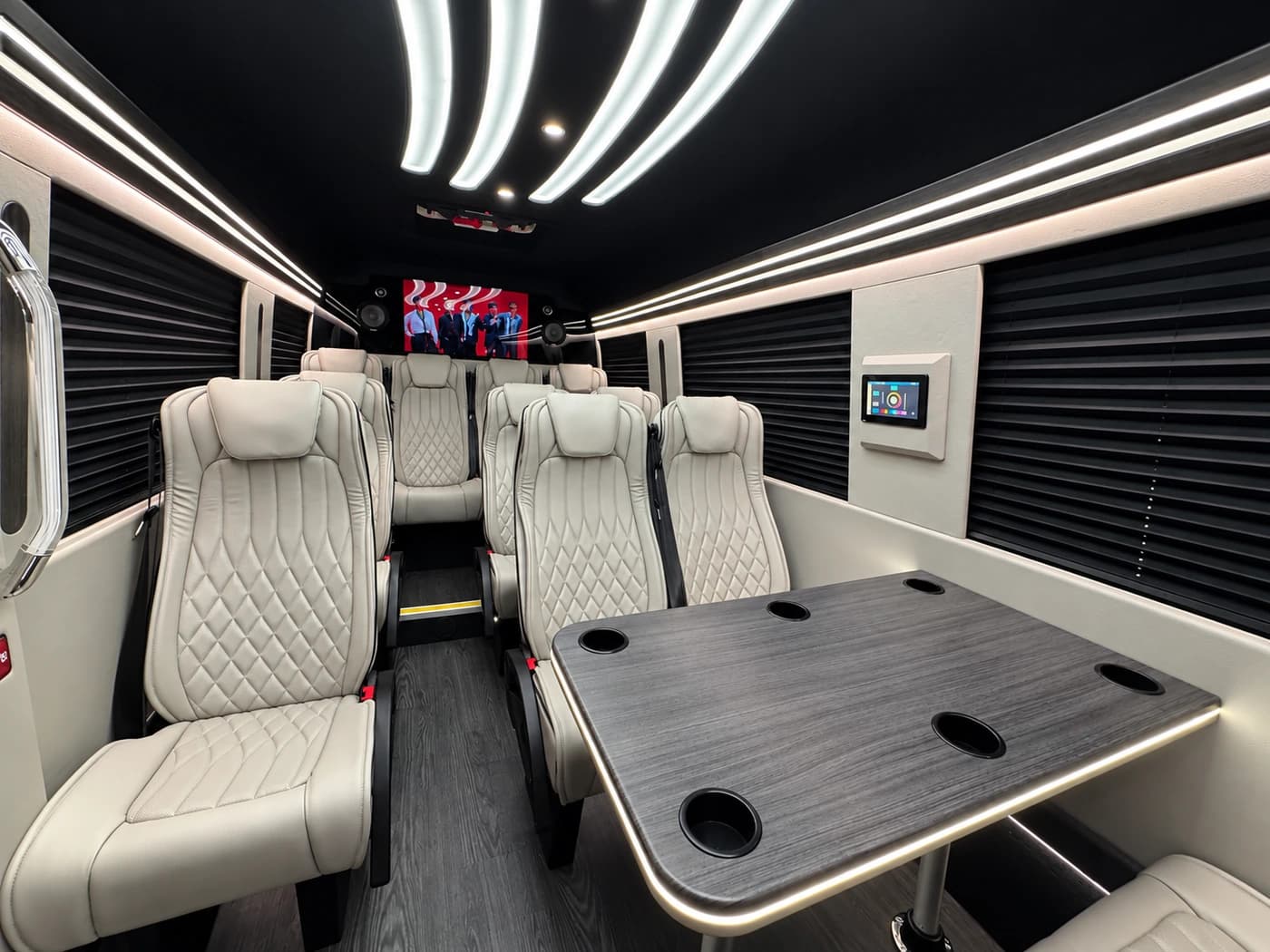 Mercedes-Benz Executive Sprinter cabin with white leather seats, a dark table, curved ceiling lights and screens.